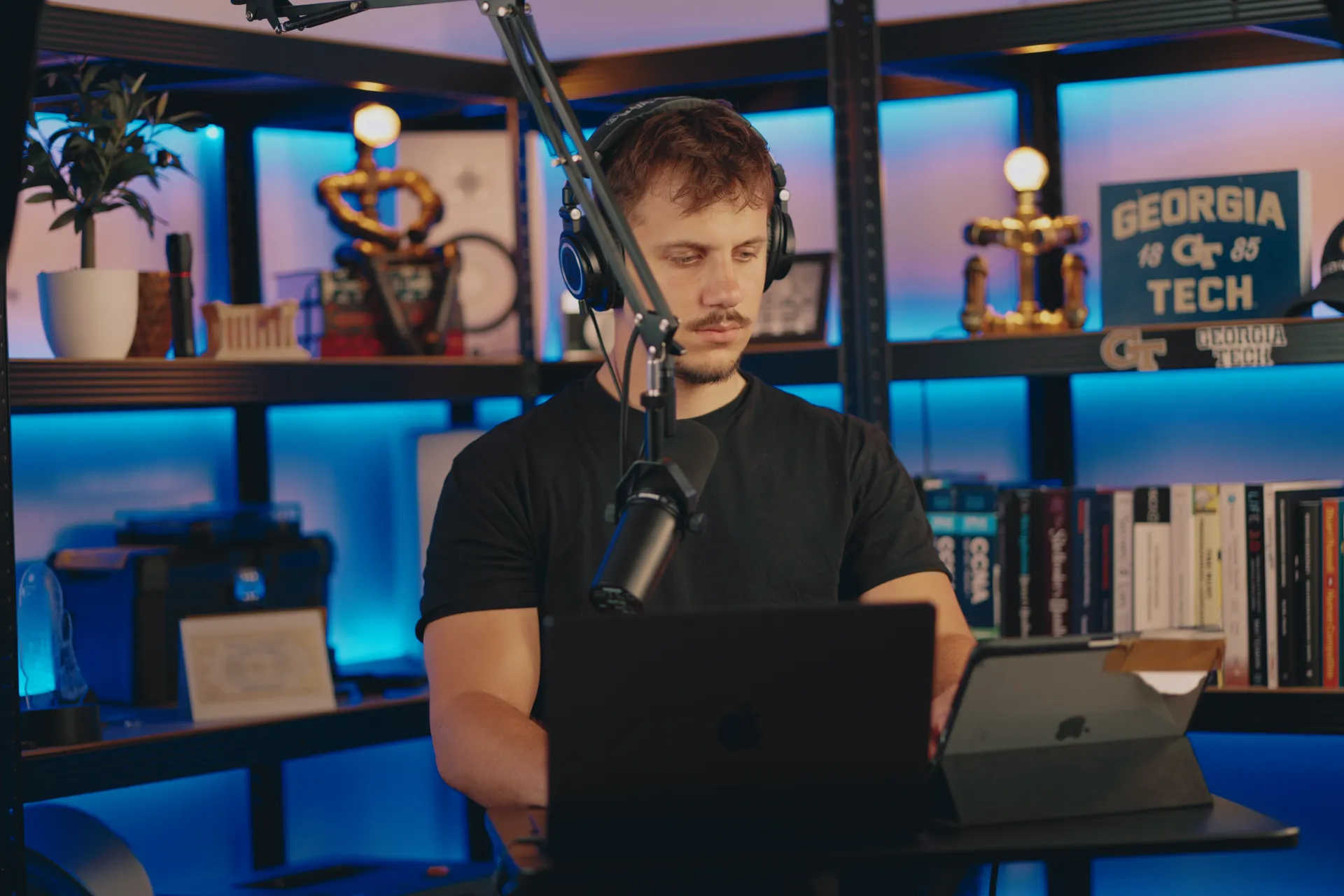 Dimitrios Prasakis, systems security researcher, working at his desk wearing headphones with a microphone and laptop, blue-lit shelves with Georgia Tech memorabilia in the background